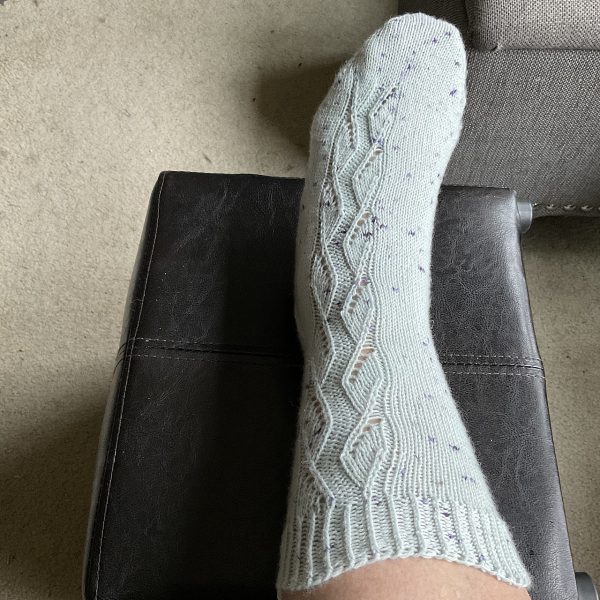 A sock handknit in white speckled yarn showing the zigzag lace pattern up the outside of the foot and leg