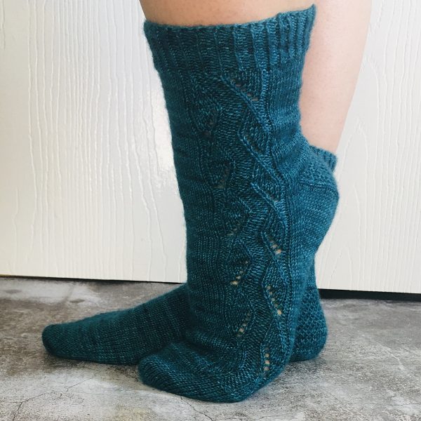 A pair of socks handknit in teal yarn showing the zigzag lace pattern up the outside of one foot and leg,