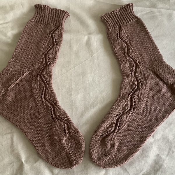 A pair of socks handknit in brown yarn laid flat to show the zigzag lace pattern up the outside of each foot and leg