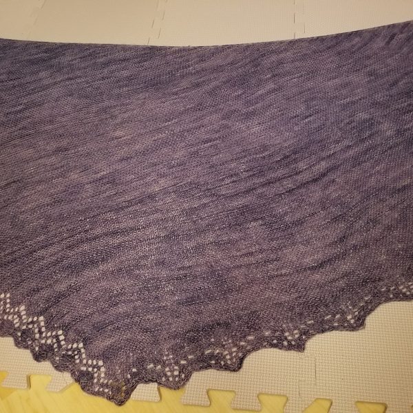 A purple shawl with a diamond edge and a rippled border folded on a blocking mat