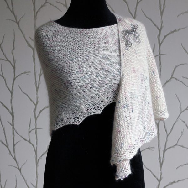 A white speckled shawl with a rippling lace pattern along the edge modelled on a mannequin with a silver shawl pin