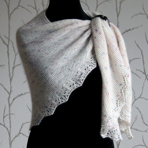 A white speckled shawl with a diamond lace pattern along one edge and a rippled border on the other modelled on a mannequin with a shawl cuff