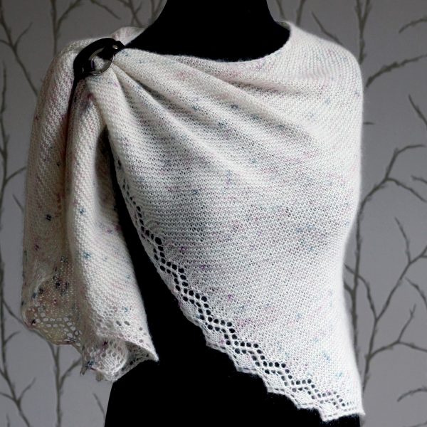 A white speckled shawl with a diamond lace pattern along the edge modelled on a mannequin with a shawl cuff