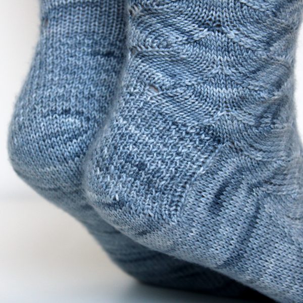 A modelled pair of socks knit in light blue grey yarn showing the textured heel pattern