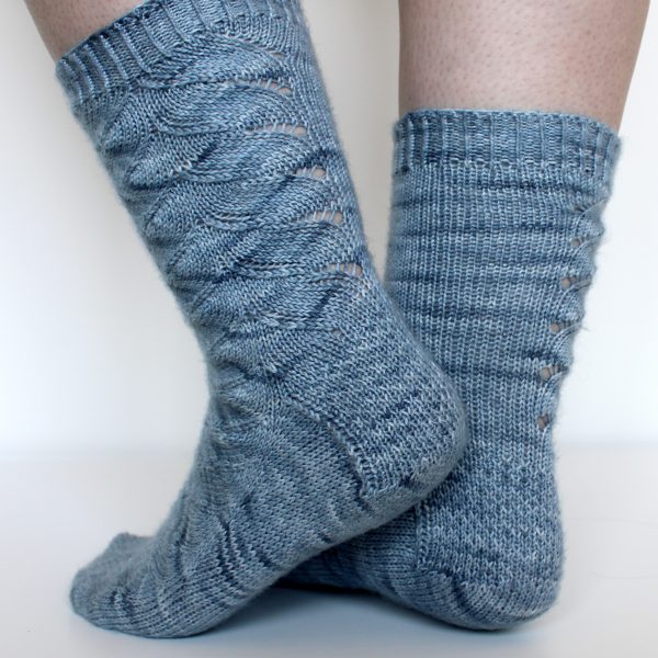 A modelled pair of socks knit in light blue grey yarn with a lace pattern swirling to the outside of the foot and into the middle of the leg and showing the textured heel pattern