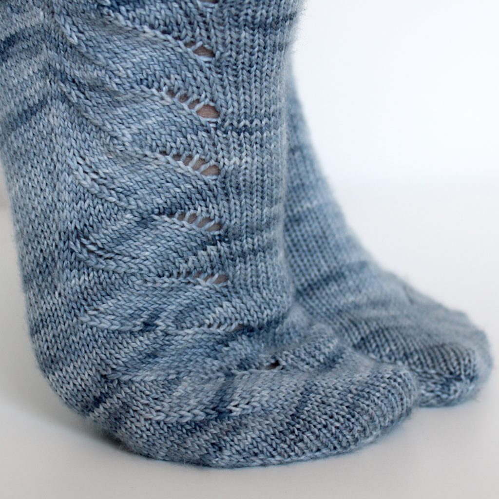 A modelled pair of socks knit in light blue grey yarn with a lace pattern swirling to the outside of the foot