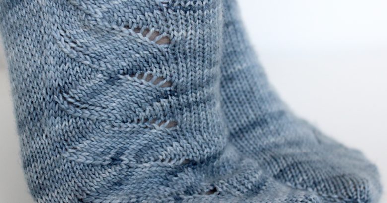 A modelled pair of socks knit in light blue grey yarn with a lace pattern swirling to the outside of the foot