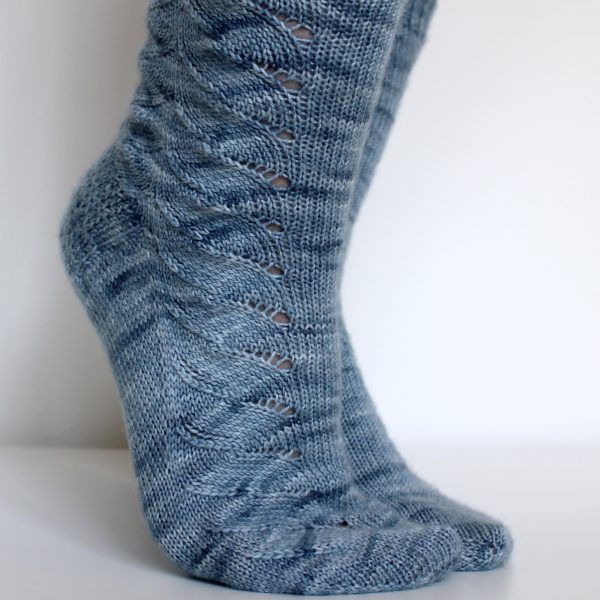 A modelled pair of socks knit in light blue grey yarn with a lace pattern swirling to the outside of the foot and into the middle of the leg