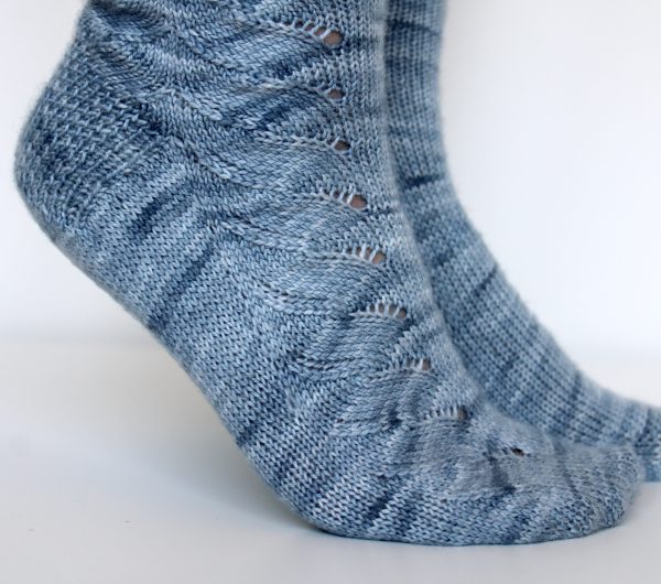 A modelled pair of socks knit in light blue grey yarn with a lace pattern swirling to the outside of the foot and into the middle of the leg