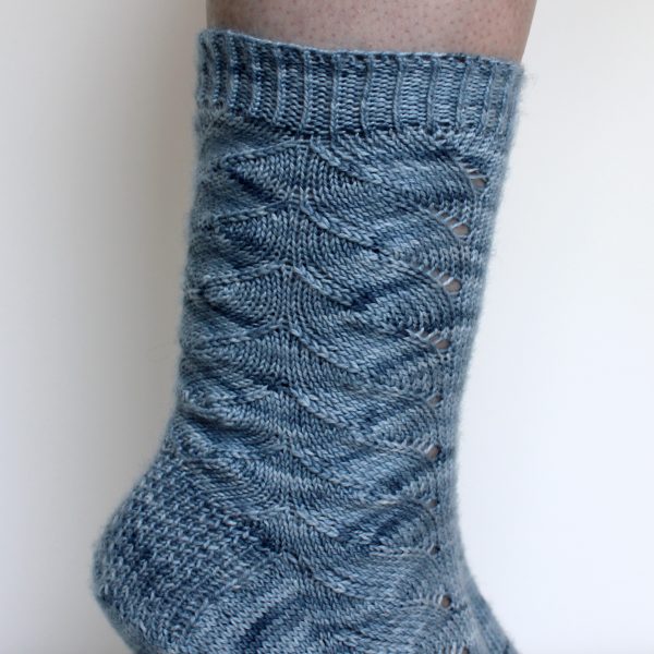 A modelled pair of socks knit in light blue grey yarn with a lace pattern swirling to the outside of the foot and into the middle of the leg