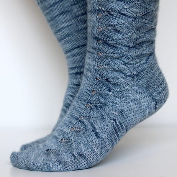 A modelled pair of socks knit in light blue grey yarn with a lace pattern swirling to the outside of the foot and into the middle of the leg