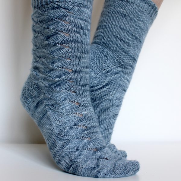 A modelled pair of socks knit in light blue grey yarn with a lace pattern swirling to the outside of the foot and into the middle of the leg