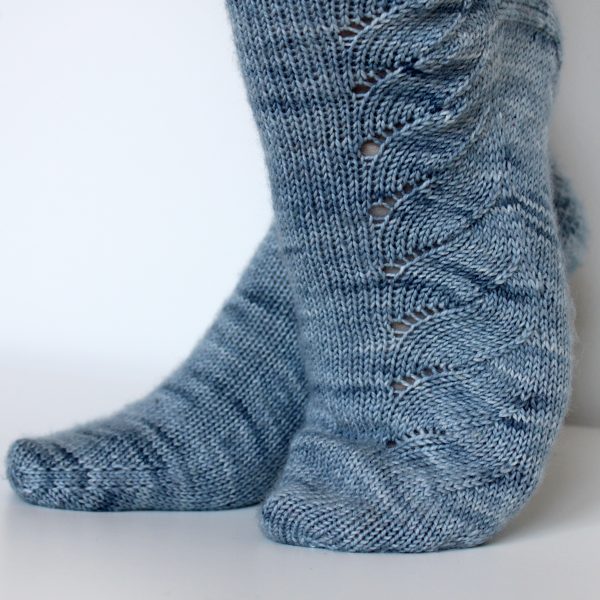 A modelled pair of socks knit in light blue grey yarn with a lace pattern swirling to the outside of the foot