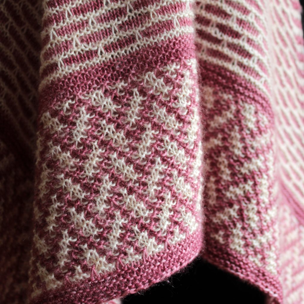 A close up on the border of a mosaic knit shawl. It is knit in white and red yarn with a brickwork pattern on the body and herringbone pattern border, displayed on a black mannequin.