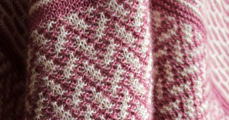 A close up on the border of a mosaic knit shawl. It is knit in white and red yarn with a brickwork pattern on the body and herringbone pattern border, displayed on a black mannequin.