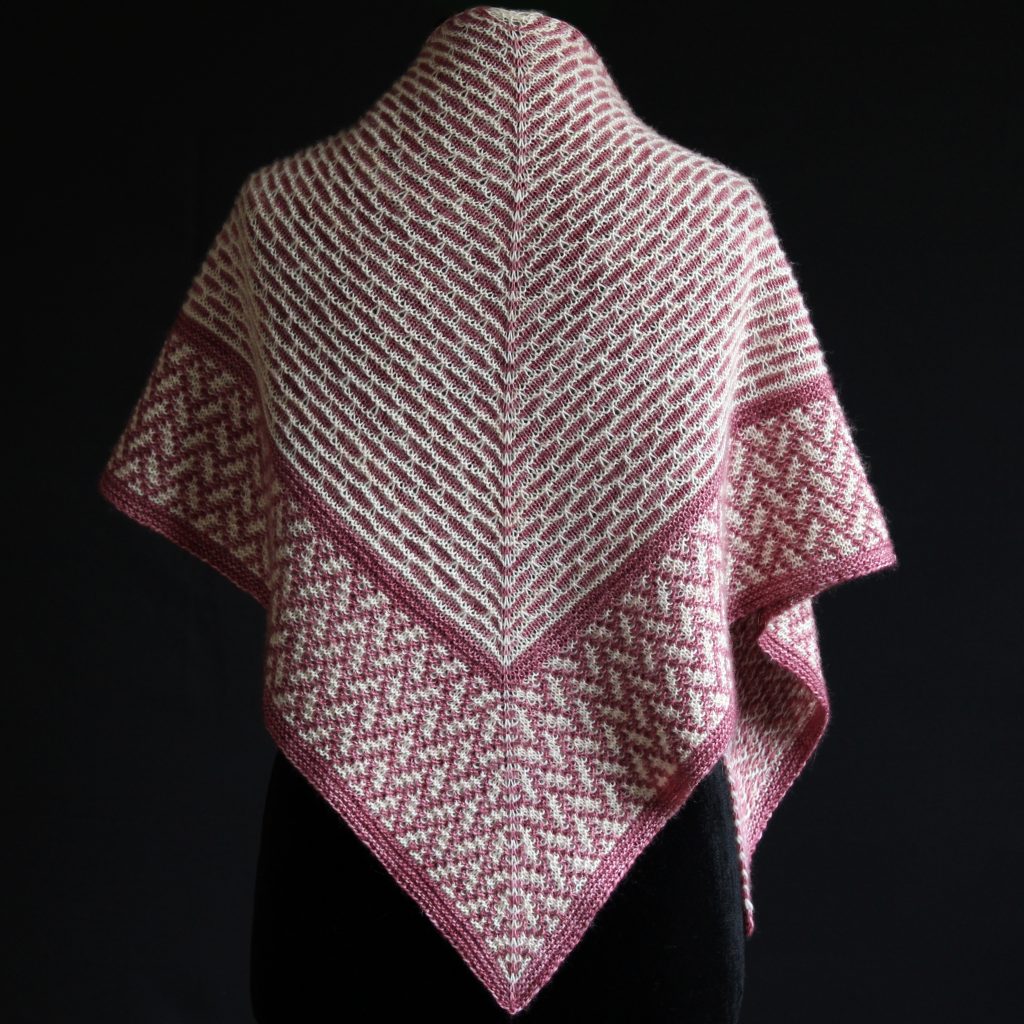 A mosaic knit shawl knit in white and red yarn with a brickwork pattern on the body and herringbone pattern border, displayed on a black mannequin.