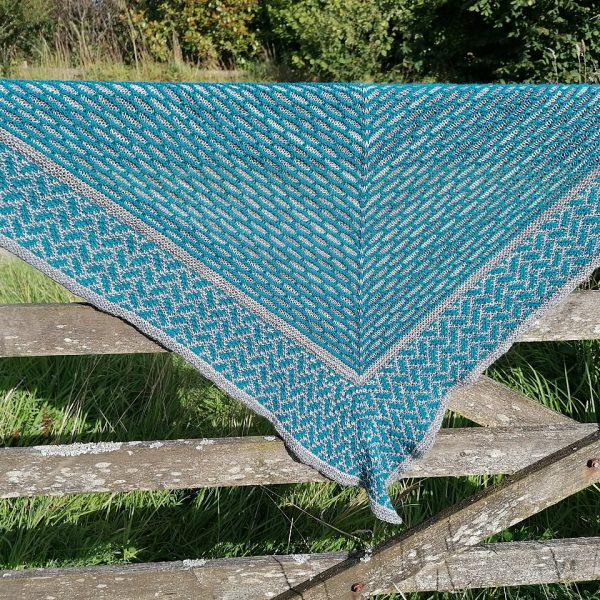 A mosaic knit shawl knit in grey and teal yarn with a brickwork pattern on the body and herringbone pattern border, displayed over a gate