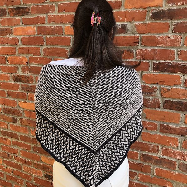 A mosaic knit shawl knit in white and black yarn with a brickwork pattern on the body and herringbone pattern border, worn over a woman's shoulders from behind
