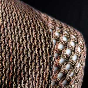 A close up on the shoulder of a cowlette knit in brown yarn with a textured slipped stitch pattern on the body and a wide lace border