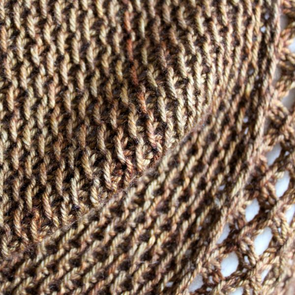 A close up on the textured pattern of a cowlette knit in brown yarn with a textured slipped stitch pattern on the body and a wide lace border