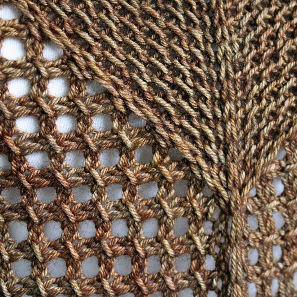 A close up on the spine of a cowlette knit in brown yarn with a textured slipped stitch pattern on the body and a wide lace border