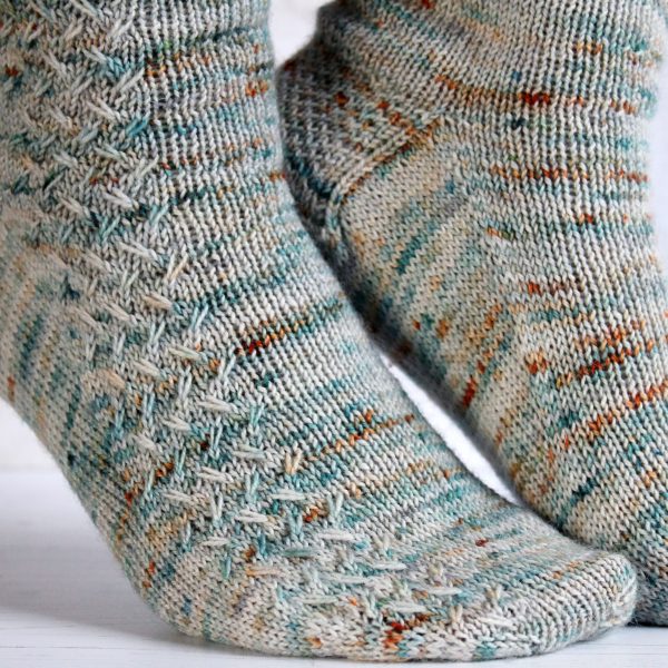 A modelled pair of socks with a rippling cable pattern up the side, knit in grey yarn with blue, orange and white speckles