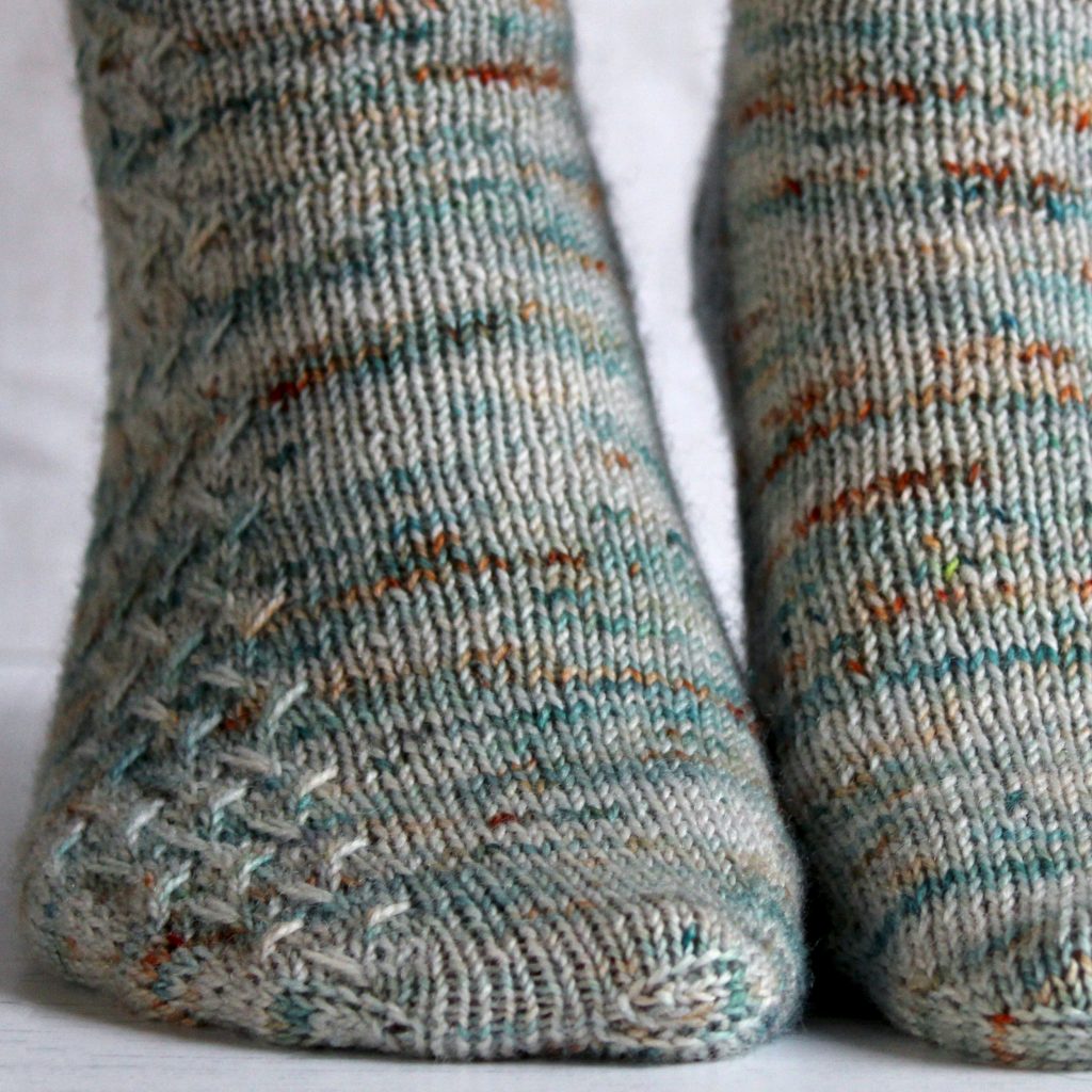 A modelled pair of socks with a rippling cable pattern up the side, knit in grey yarn with blue, orange and white speckles