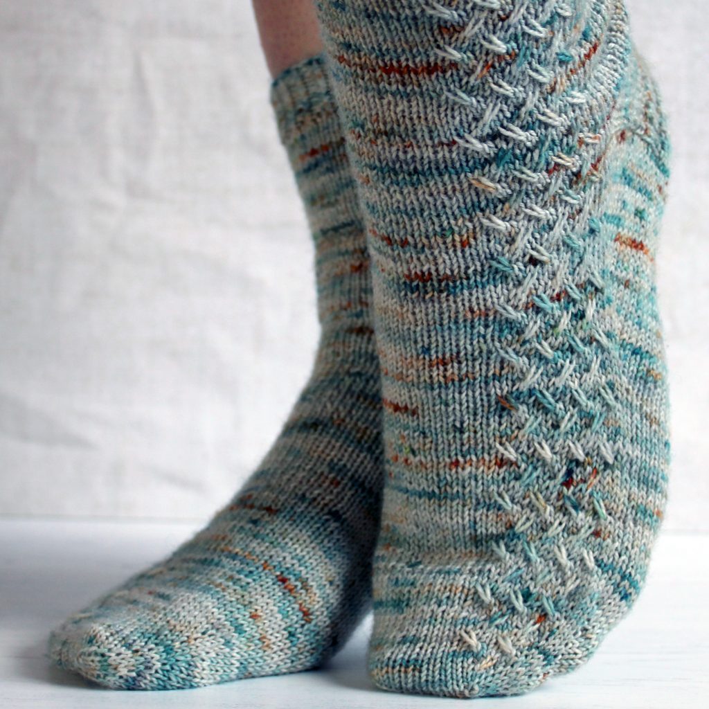 A modelled pair of socks with a rippling cable pattern up the side, knit in grey yarn with blue, orange and white speckles