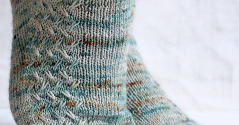 A modelled pair of socks with a rippling cable pattern up the side, knit in grey yarn with blue, orange and white speckles