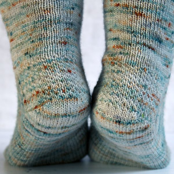A modelled pair of socks showing the textured heel pattern, knit in grey yarn with blue, orange and white speckles