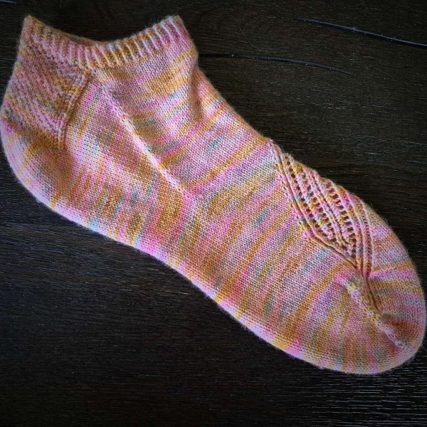 A side view of a trainer sock with a lace and twisted rib pattern travelling across the sock. The sock is knit in a pastel variegated yarn.