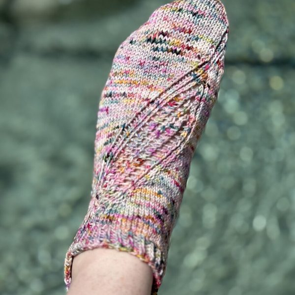 A trainer sock with a lace and twisted rib pattern travelling across the sock. The socks are knit in a colourful yarn with speckles.