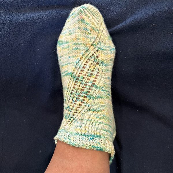 A trainer sock with a lace and twisted rib pattern travelling across the sock. The socks are knit in a white yarn with speckles.