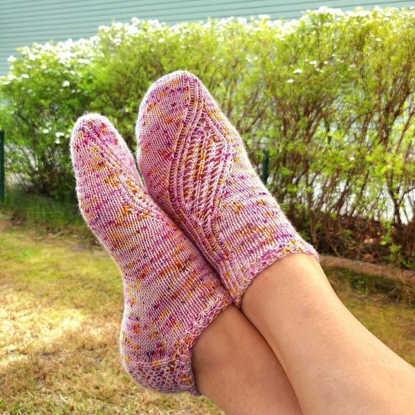 A pair of trainer socks with a lace and twisted rib pattern travelling across the sock. The socks are knit in pink yarn with speckles.