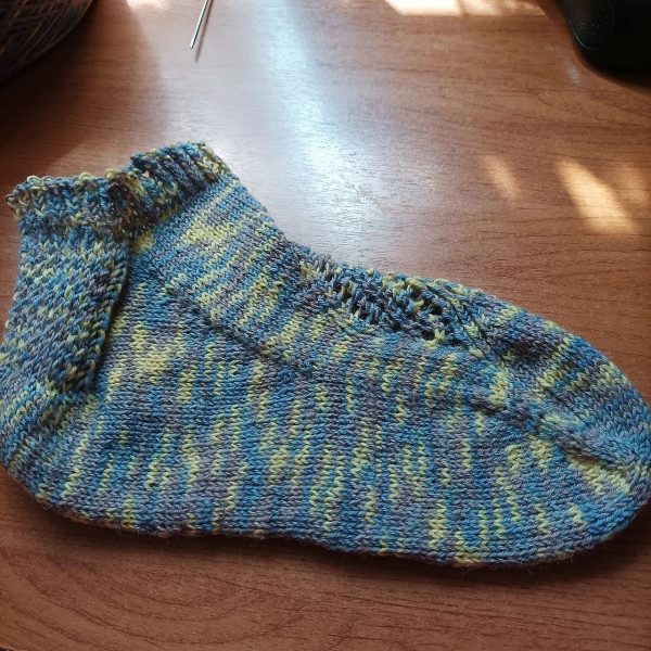 A side view of a trainer sock with a lace and twisted rib pattern travelling across the sock. The sock is knit in a blue variegated yarn.