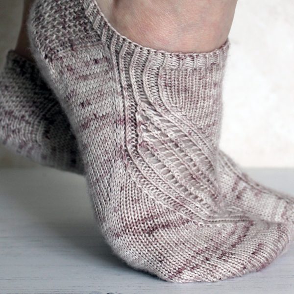 A side view of trainer socks with a lace and twisted rib pattern travelling across the sock. The socks are knit in light brown yarn with speckles.