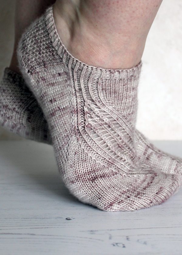 A side view of trainer socks with a lace and twisted rib pattern travelling across the sock. The socks are knit in light brown yarn with speckles.