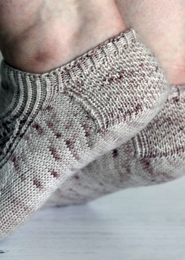 A side view textured heel of a pair of trainer socks with a lace and twisted rib pattern travelling across the sock. The socks are knit in light brown yarn with speckles.