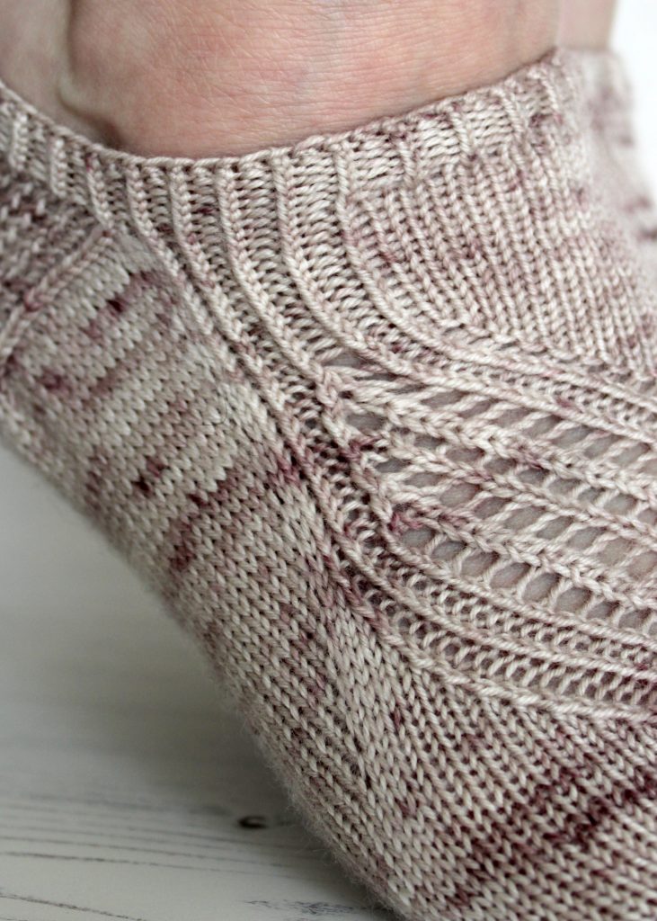 A close up on trainer socks with a lace and twisted rib pattern travelling across the sock. The socks are knit in light brown yarn with speckles.