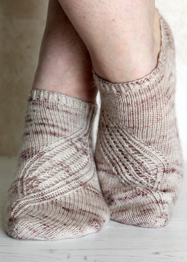 A pair of trainer socks with a lace and twisted rib pattern travelling across the sock. The socks are knit in light brown yarn with speckles.