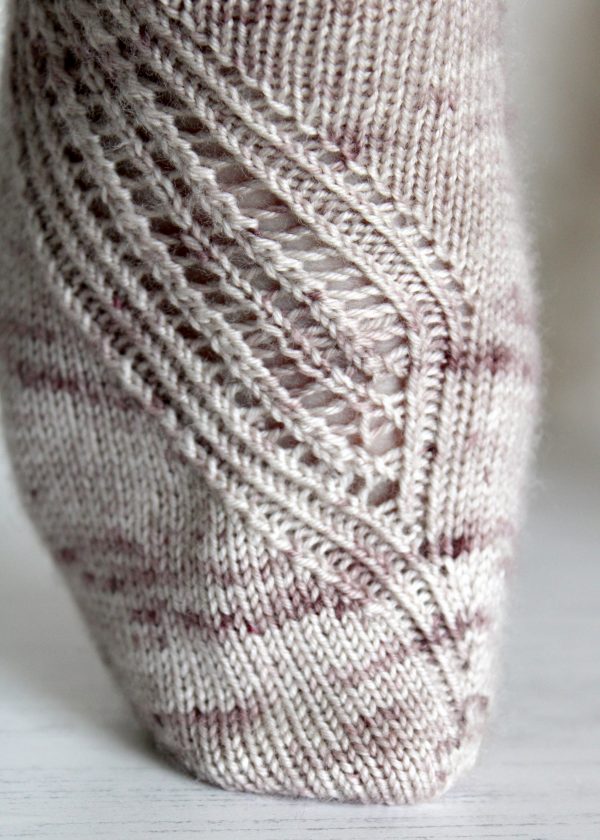 A close up on the toe of a sock with a lace and twisted rib pattern travelling across the sock. The socks are knit in light brown yarn with speckles.