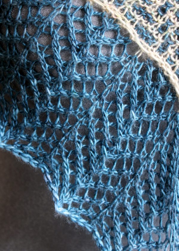 A close up on a cowlette with a mosaic body, knit in blue and white yarn, with a wide lace border knit in blue.