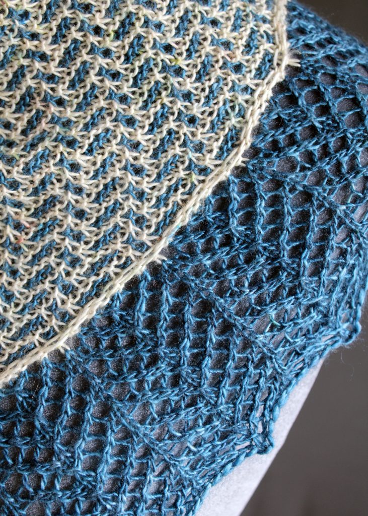 A close up on a cowlette with a mosaic body, knit in blue and white yarn, with a wide lace border knit in blue.