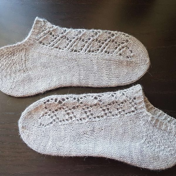 A pair of short socks featuring a lace leaf column running up the outside of the foot, knit in neutral yarn.