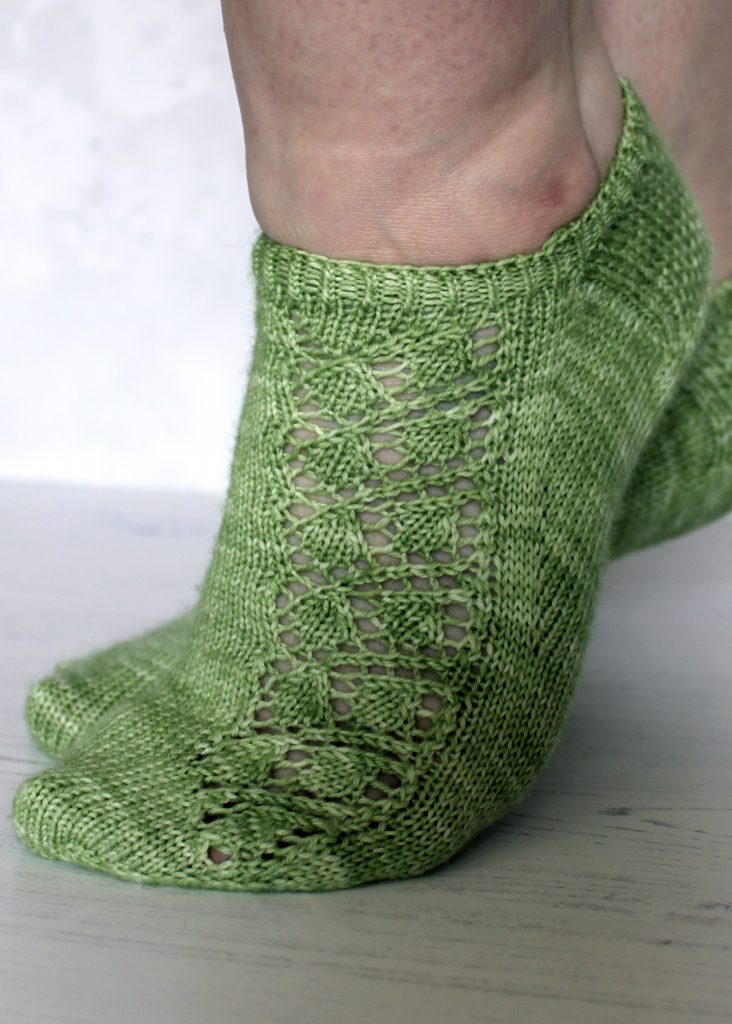A pair of modelled short socks featuring a lace leaf column running up the outside of the foot, knit in green yarn.