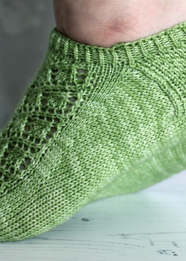 A close up on the side of a short socks howing a lace leaf pattern on the outside of the foot and the textured heel pattern, knit in green yarn.