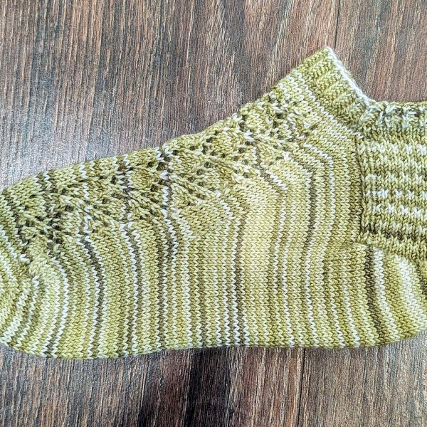A short sock featuring a lace leaf column running up the outside of the foot, knit in green striped yarn.