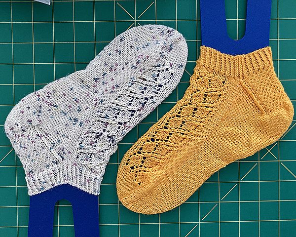 A two left short socks featuring a lace leaf column running up the outside of the foot, one knit in a yellow yarn and the other in a white speckled yarn .