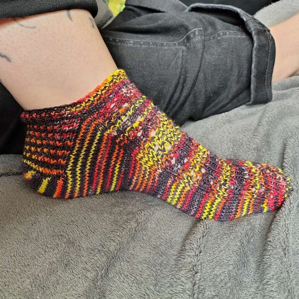 One modelled short (trainer) socks with a twisted rib and lace pattern covering the top of the foot. The socks are knit in self striping yellow, red and black yarn.