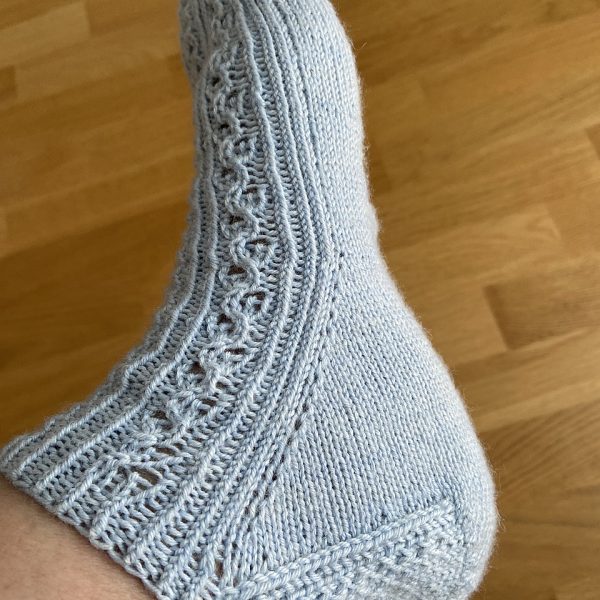 One modelled short (trainer) sock with a twisted rib and lace pattern covering the top of the foot. The sock is knit in light blue yarn.